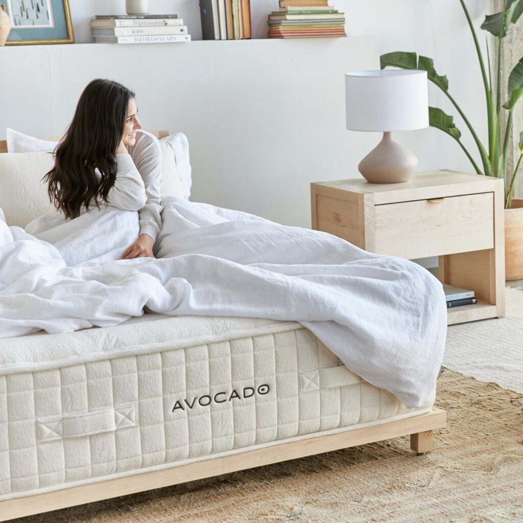 A woman is sitting and smiling on the Avocado mattress in a well-lit bedroom.